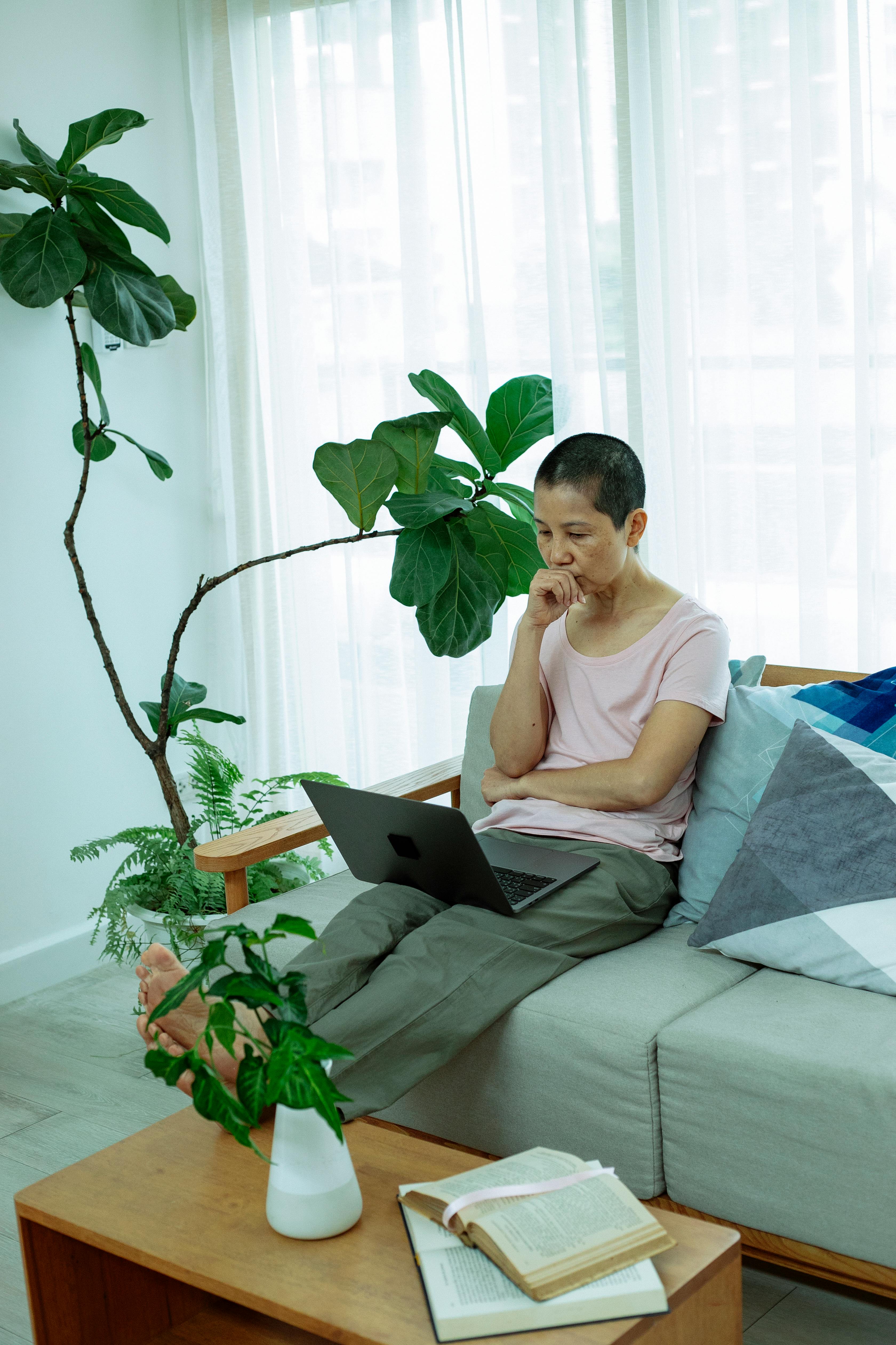 pensive ethnic female sitting on couch and watching video on laptop