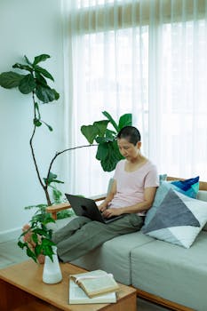 Serious ethnic self employed woman with dark short hair in casual clothes sitting on comfortable sofa and typing on laptop keyboard during remote work at home