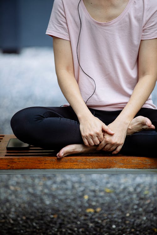 Crop faceless barefooted lady in sportswear practicing Padmasana pose while sitting on bench during yoga practice in park