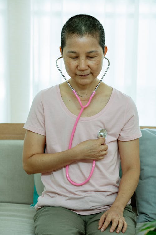 Middle aged Asian woman with short dark hair in casual clothes sitting on couch and listening heartbeat with stethoscope during recovery at home