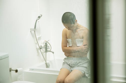 Through glass of upset Asian female with amputated breasts siting on bathtub with crossed arms and looking down before taking bath