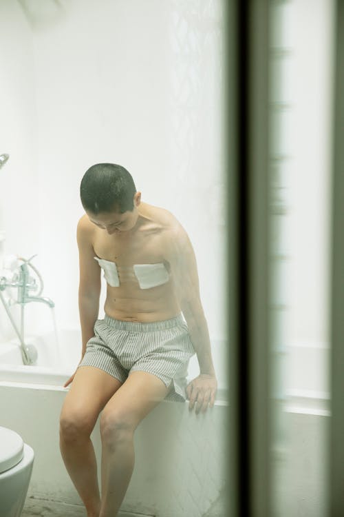 Through glass of depressed topless ethnic female with short hair and gauze bandage on amputated breasts sitting on bathtub and looking down sadly