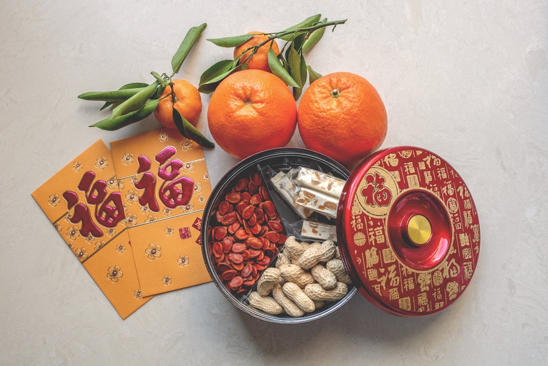 Assortment of Lunar New Year treats including nuts, seeds, and candies in a festive container, accompanied by fresh oranges and lucky red envelopes