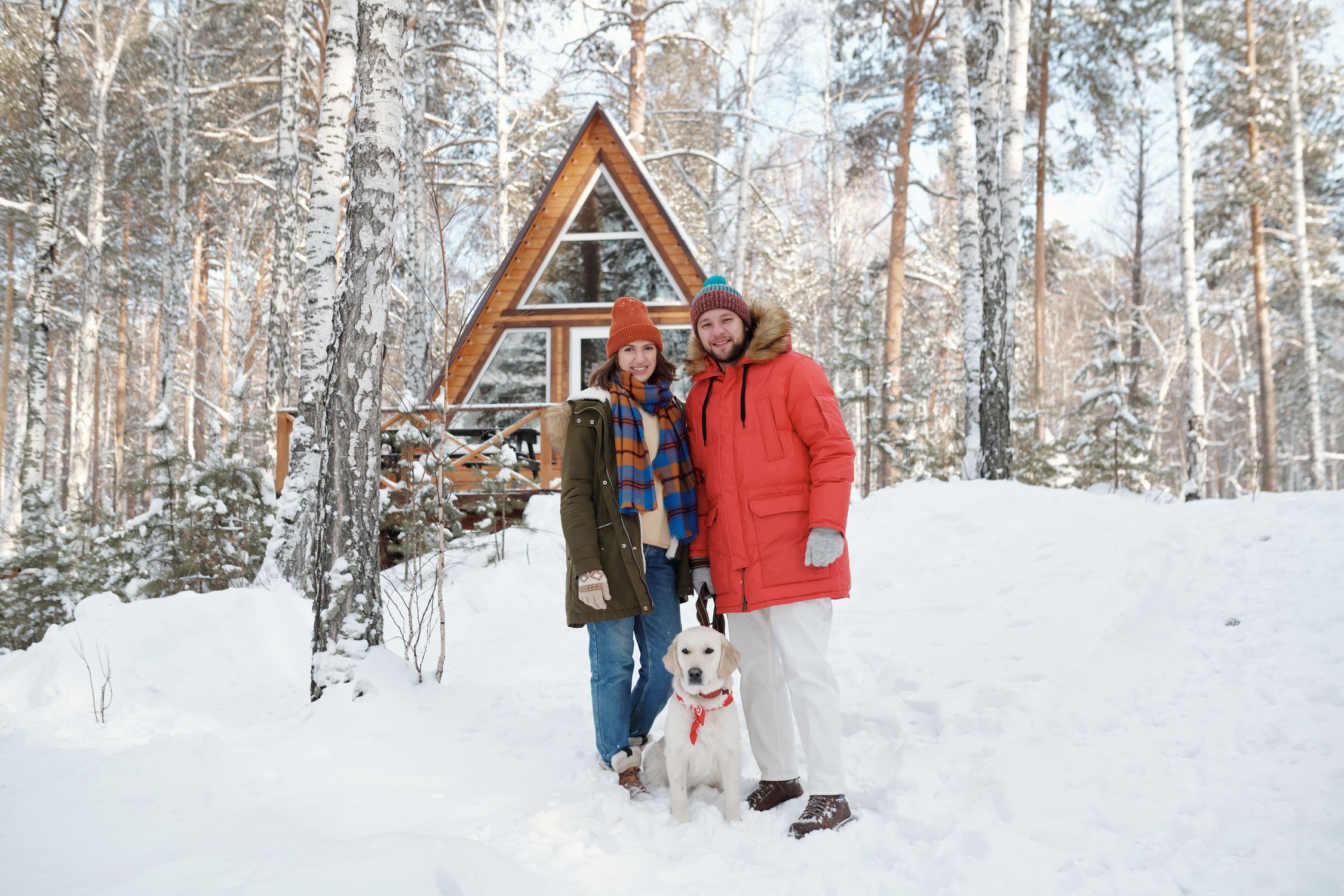 Happy couple with their dog outside a cozy winter cabin in a snowy forest