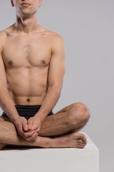 A shirtless man sitting cross-legged on a white surface, focused on relaxation and self-care.