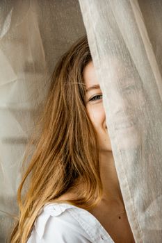 A woman with long hair smiles while partially concealed by a sheer curtain indoors.