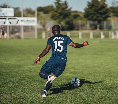 Back view unrecognizable African American football player kicking ball on outdoor grassy field during match on summer day