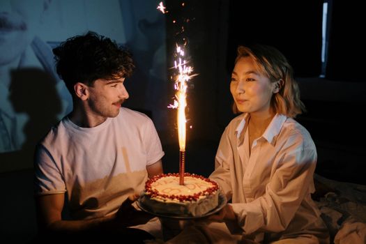 Couple celebrating a birthday with a cake and sparkling candle indoors, captured in a warm, intimate setting.