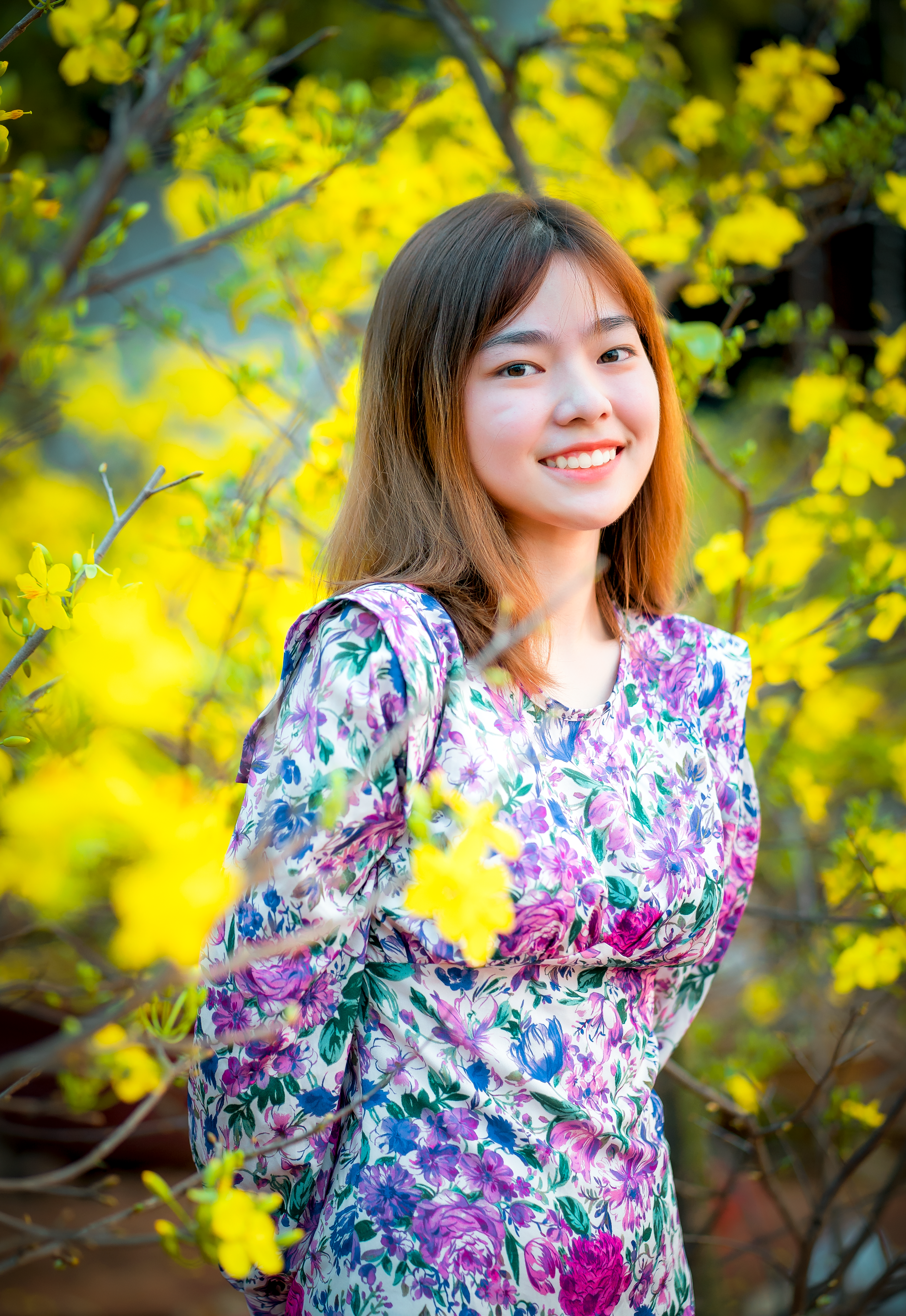 A young woman in a floral dress smiles amidst vibrant yellow blooms in an outdoor setting.