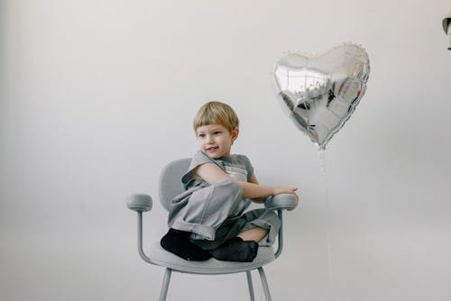 Free A Kid Sitting on a Chair Near a Silver Balloon Stock Photo