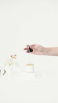 Minimalist shot of a hand holding a dropper over a skincare bottle beside a white lily.