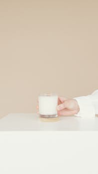 A minimalist scene featuring a hand holding a glass of milk on a white table against a beige background.
