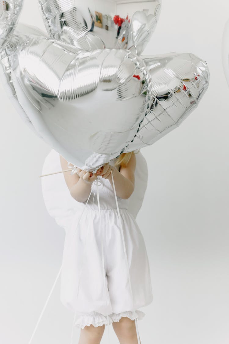 A Child In A White Dress Holding Heart Shaped Balloons