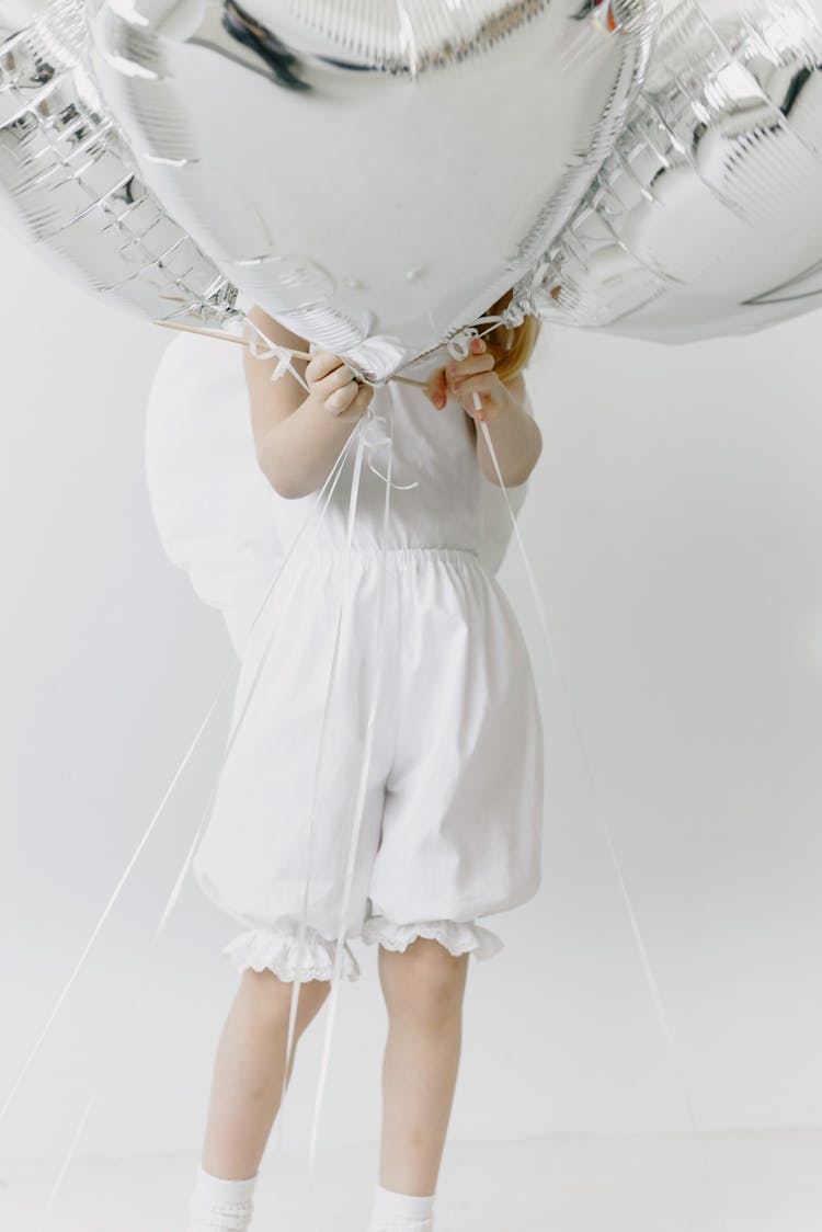 A Child In A White Dress Holding Heart Shaped Balloons