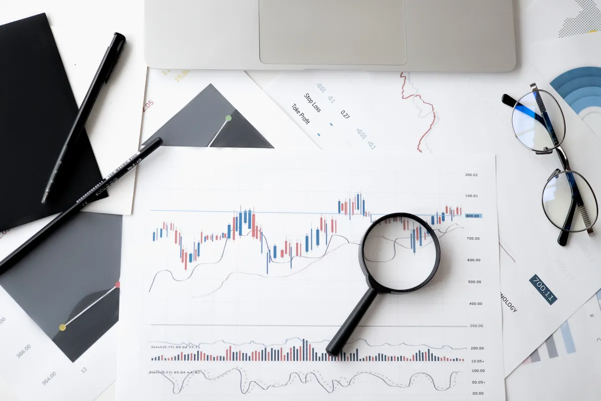 Printed candlestick charts on a desk with a magnifying glass, pen, and glasses, used to inspect backtest detail.