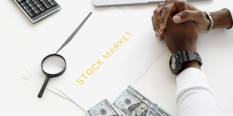 Hands folded over stock market documents with calculator and cash on desk.