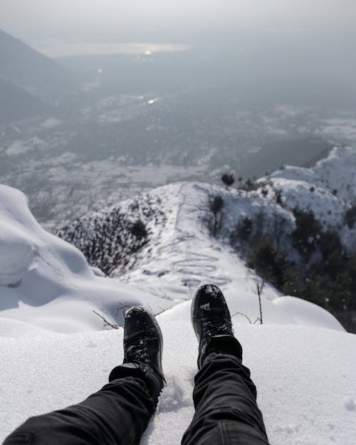 A Person in Black Pants and Shoes Sitting on a Snow Covered Ground at the Mountain