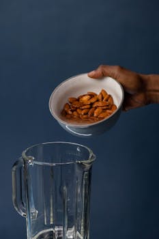 A hand pours almonds from a ceramic bowl into a glass blender against a neutral backdrop.
