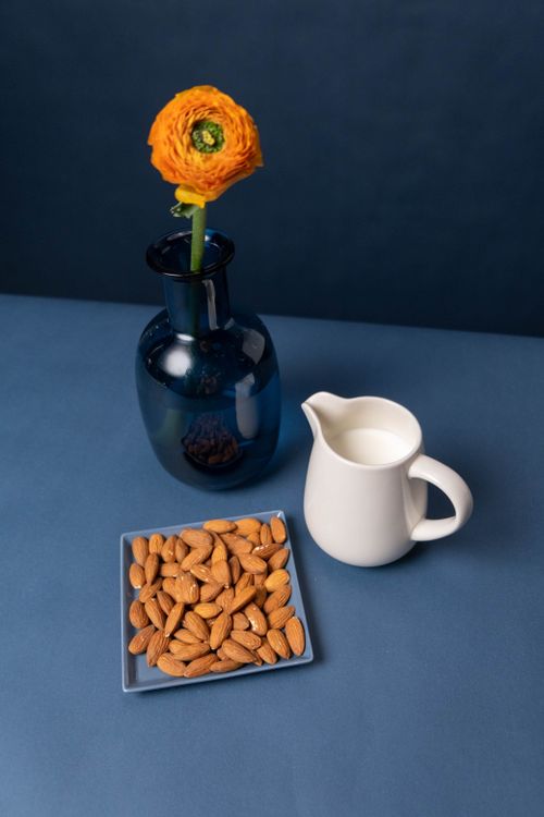 Free A minimalist still life featuring almonds, a glass vase with an orange flower, and a milk pitcher on a blue surface. Stock Photo