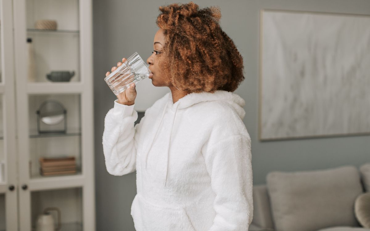 A woman in a cozy hoodie drinks water at home, promoting hydration and relaxation.