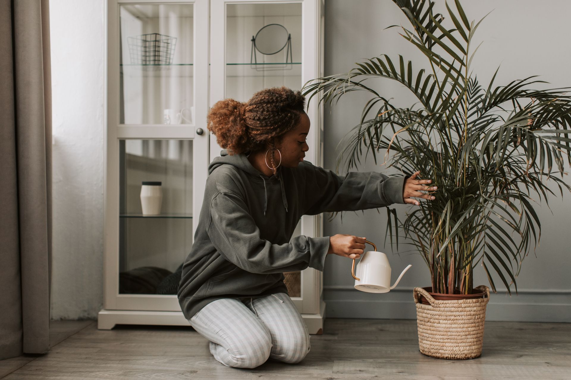 A woman kneels indoors while watering a potted palm plant with a white watering can.