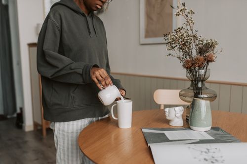 Free Woman in a gray hoodie pours coffee from a ceramic cup into a mug, with a flower vase on a wooden table. Stock Photo