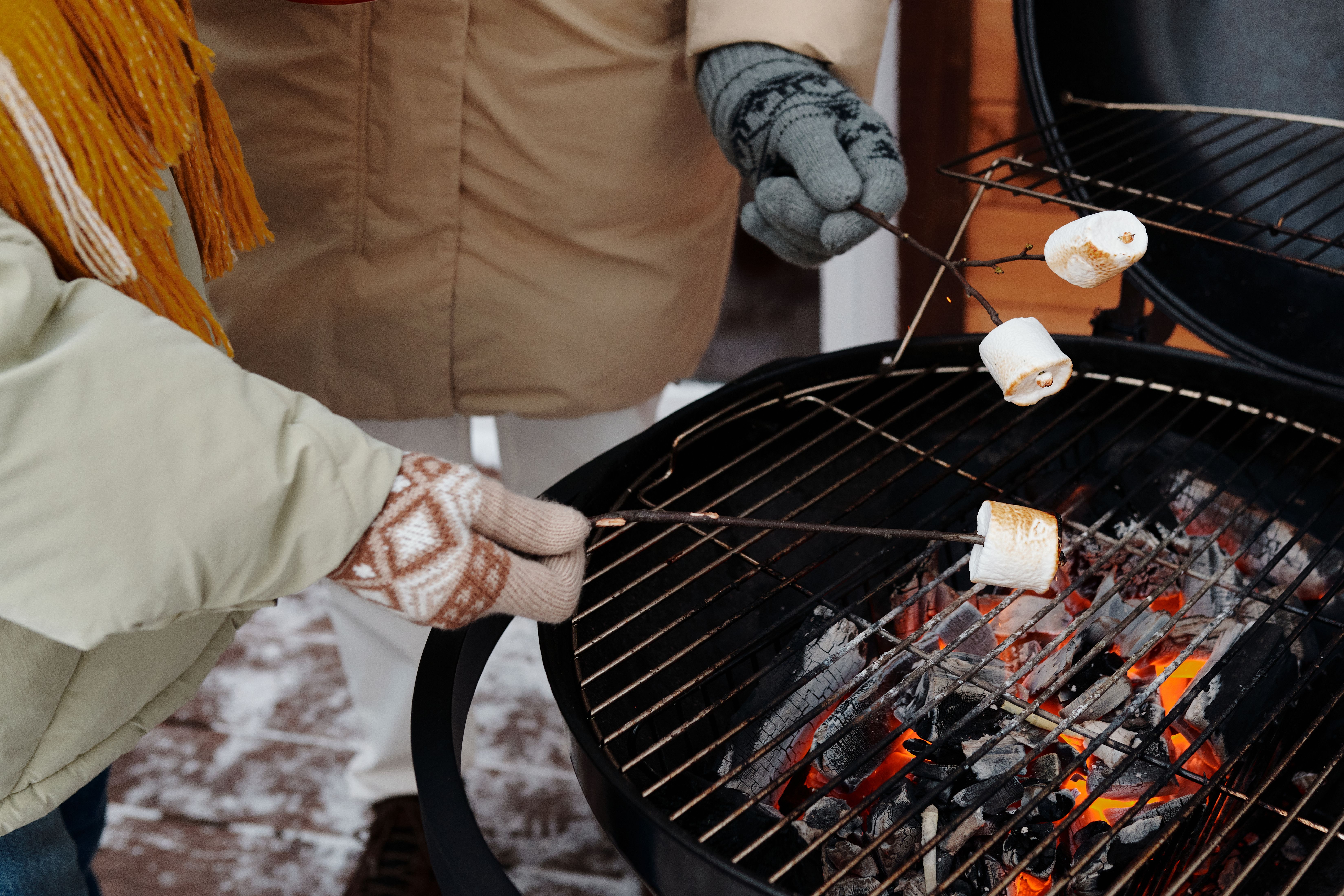 Pessoa fazendo marshmallows assados em uma churrasqueira portátil durante o inverno, com roupas quentes e luvas.
