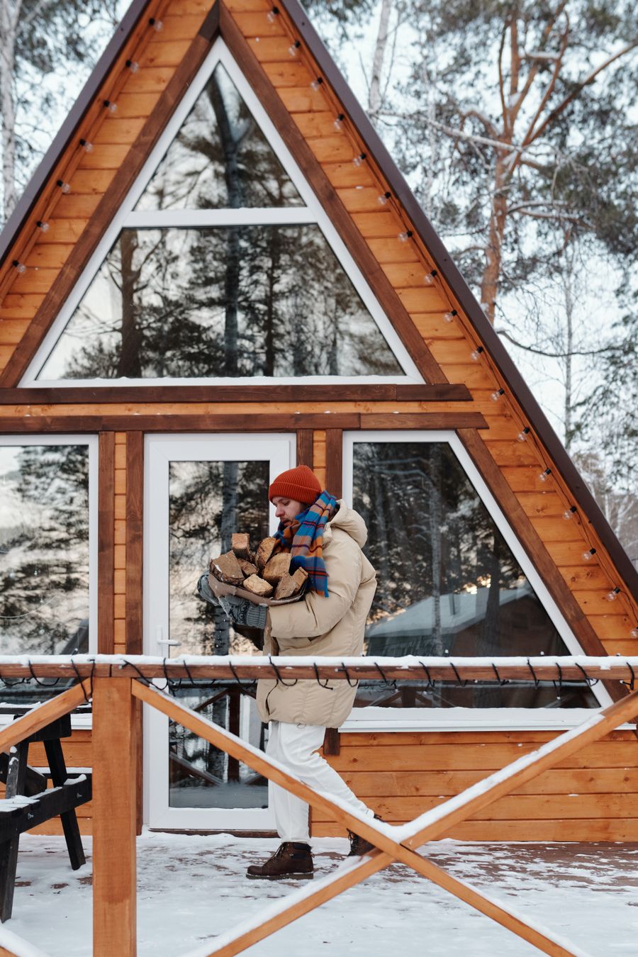 A person in a knitted hat and winter jacket carrying an armful of firewood in front of a snowy A-frame cabin