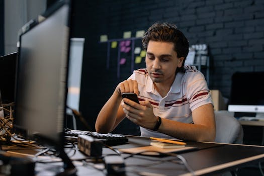 Young man in modern office using smartphone at his desk, focusing on multitasking work.