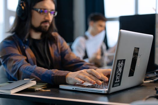 A man wearing headphones focused on typing at his laptop in a contemporary office setting.
