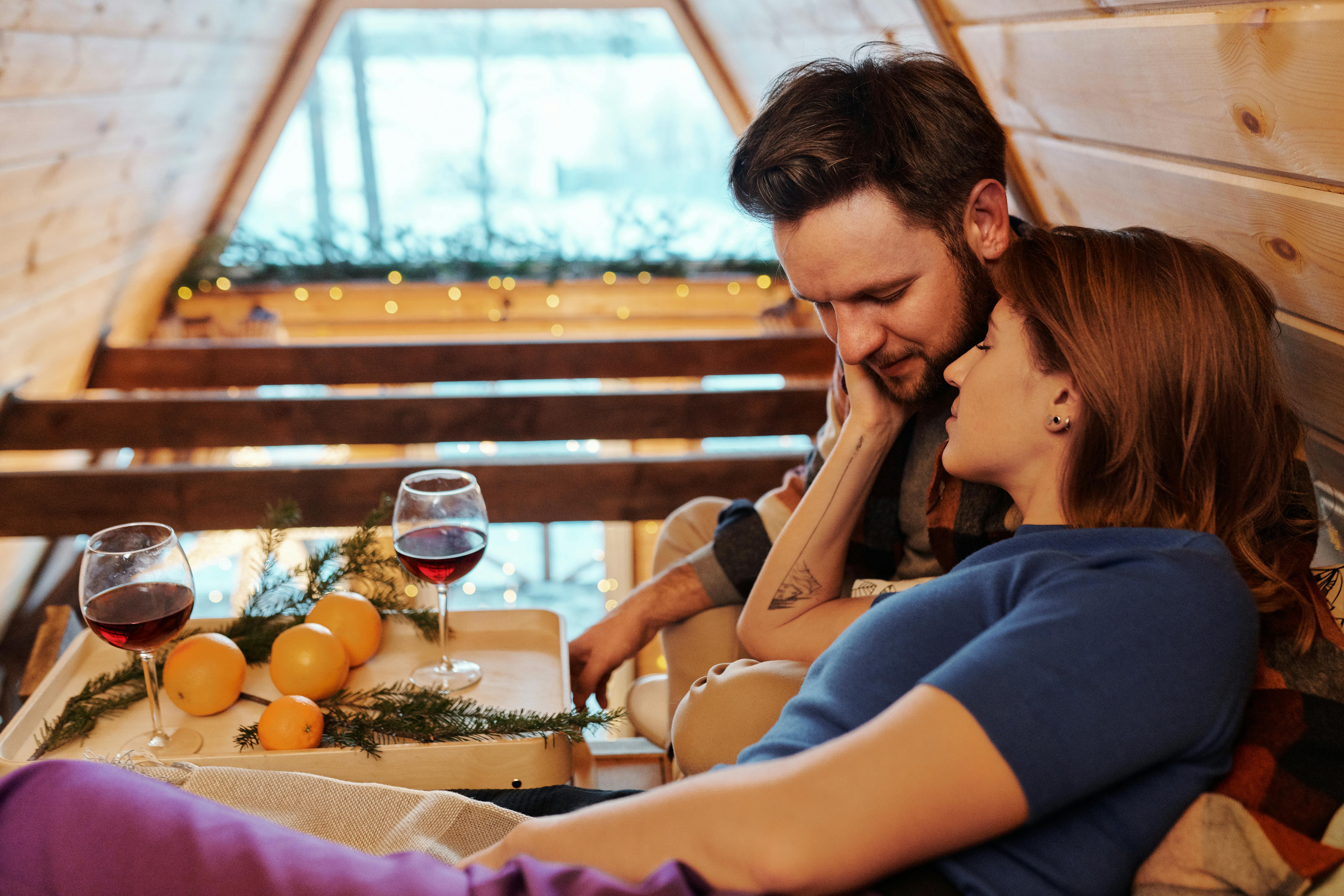 Romantic couple relaxing indoors with wine and oranges in a wooden cabin setting.
