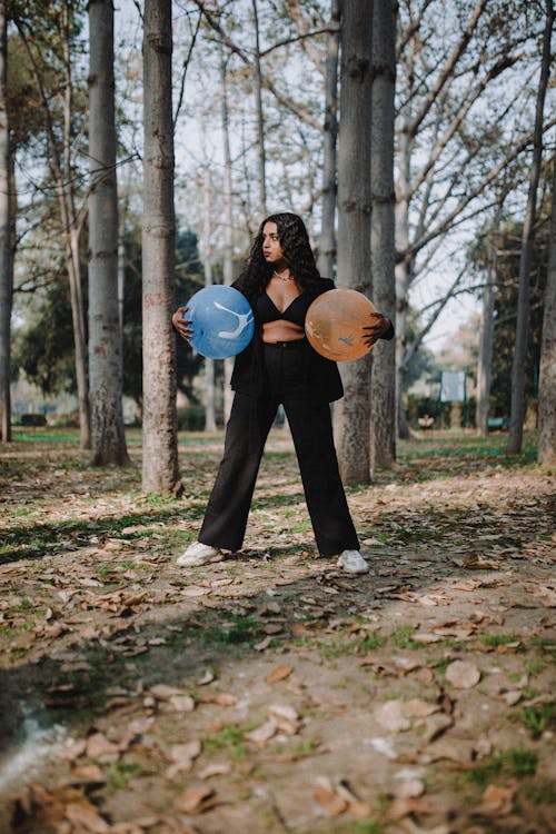 Full body of serious young female in black outfit standing with balloons while looking away in park with tall trees in autumn