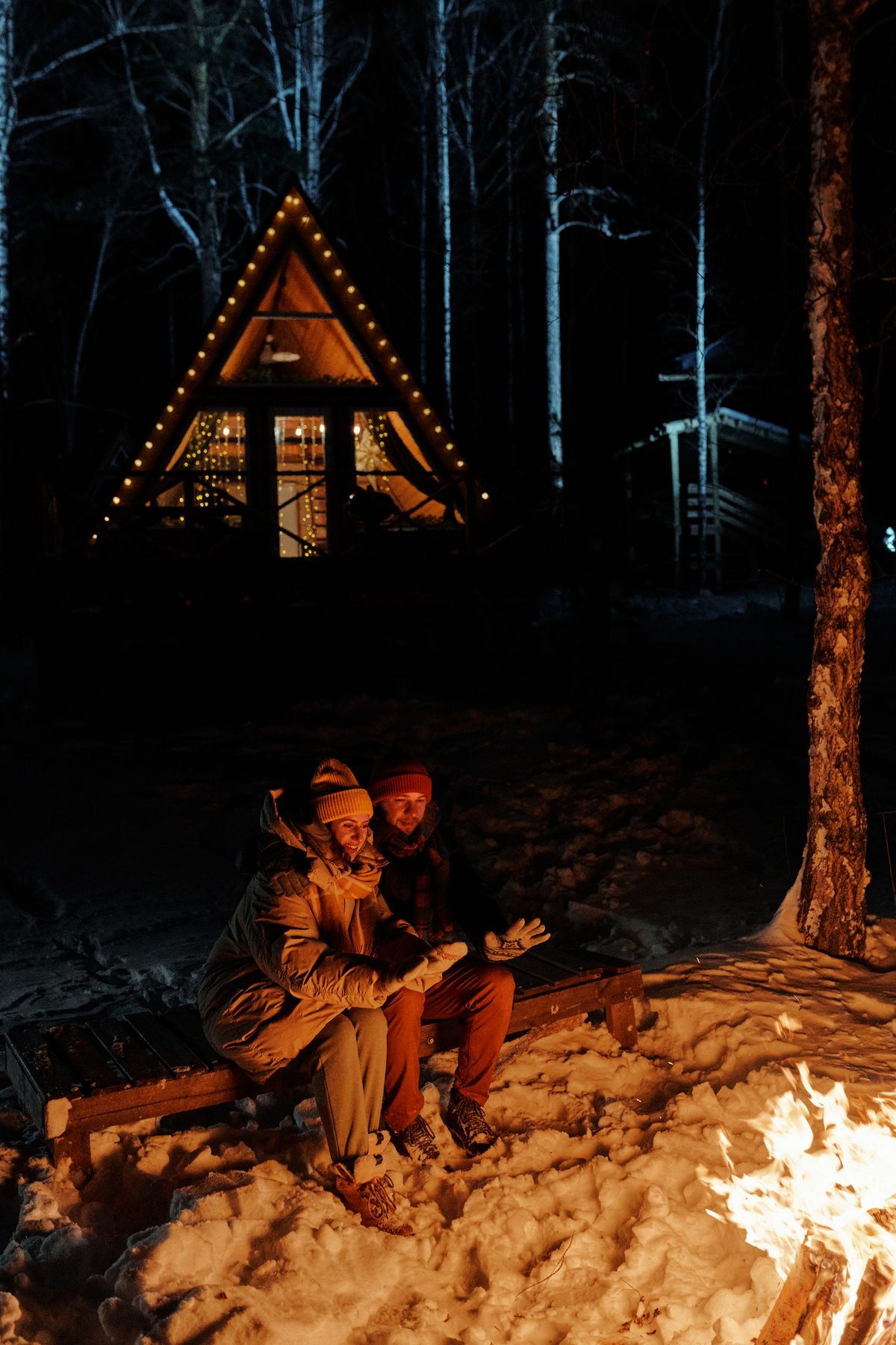 People in winter gear around a bonfire near a snowy lodge at night.