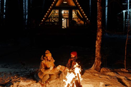 Free Couple enjoying a warm bonfire outside a cabin in snowy winter forest. Perfect cozy night scene. Stock Photo