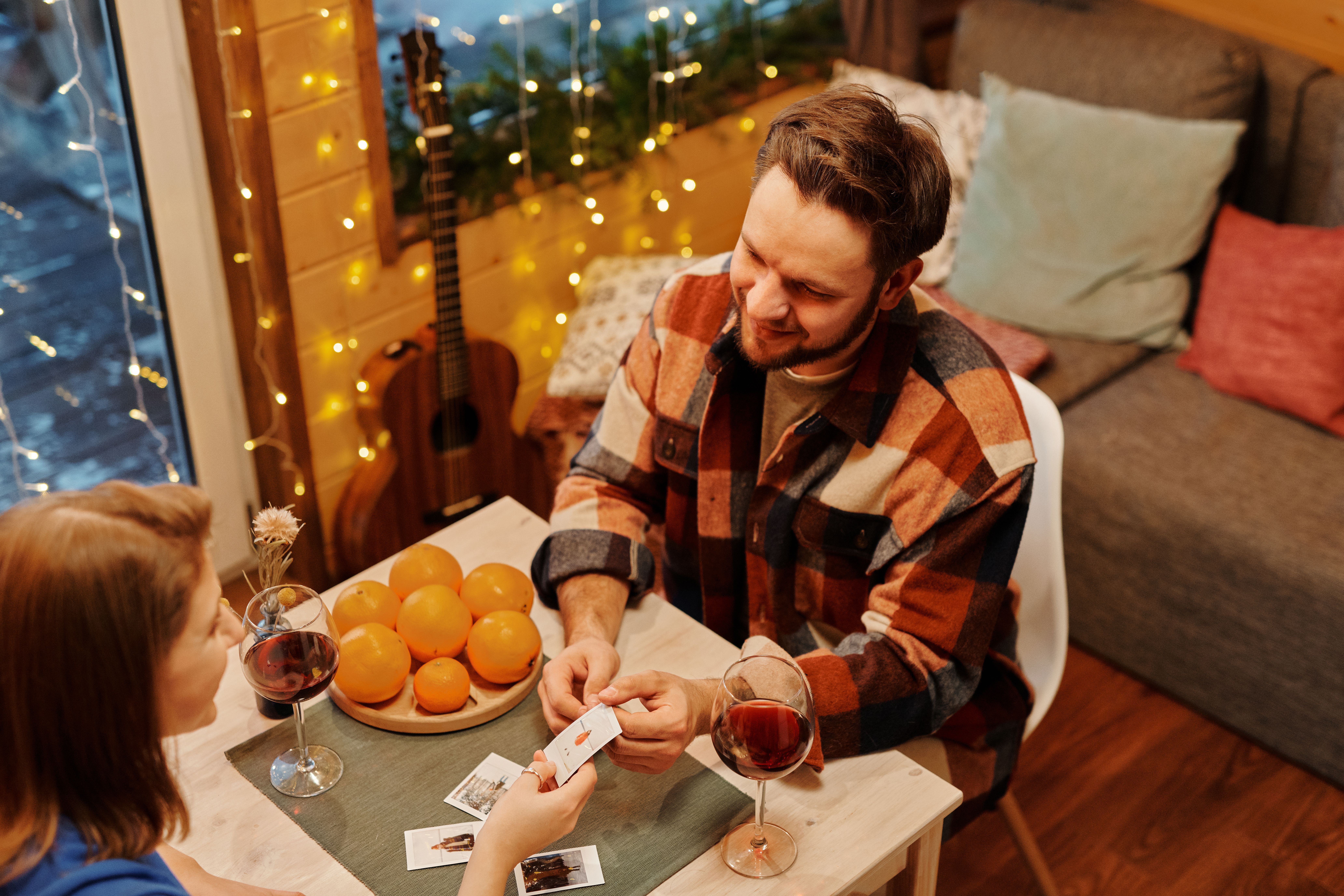 A couple enjoys a cozy indoor evening playing cards with wine and oranges on the table.