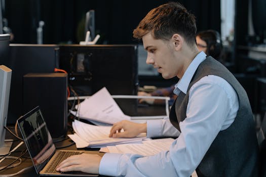 Young male professional focused on work with laptop and documents at office desk.