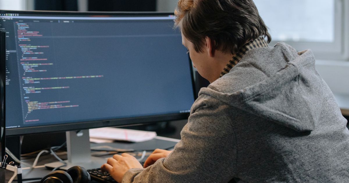 A man in a hoodie coding at a desk with large monitors in an office setting.