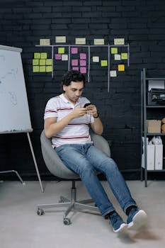 A young man in a casual office setting texting on his smartphone amidst sticky notes and a flip chart.