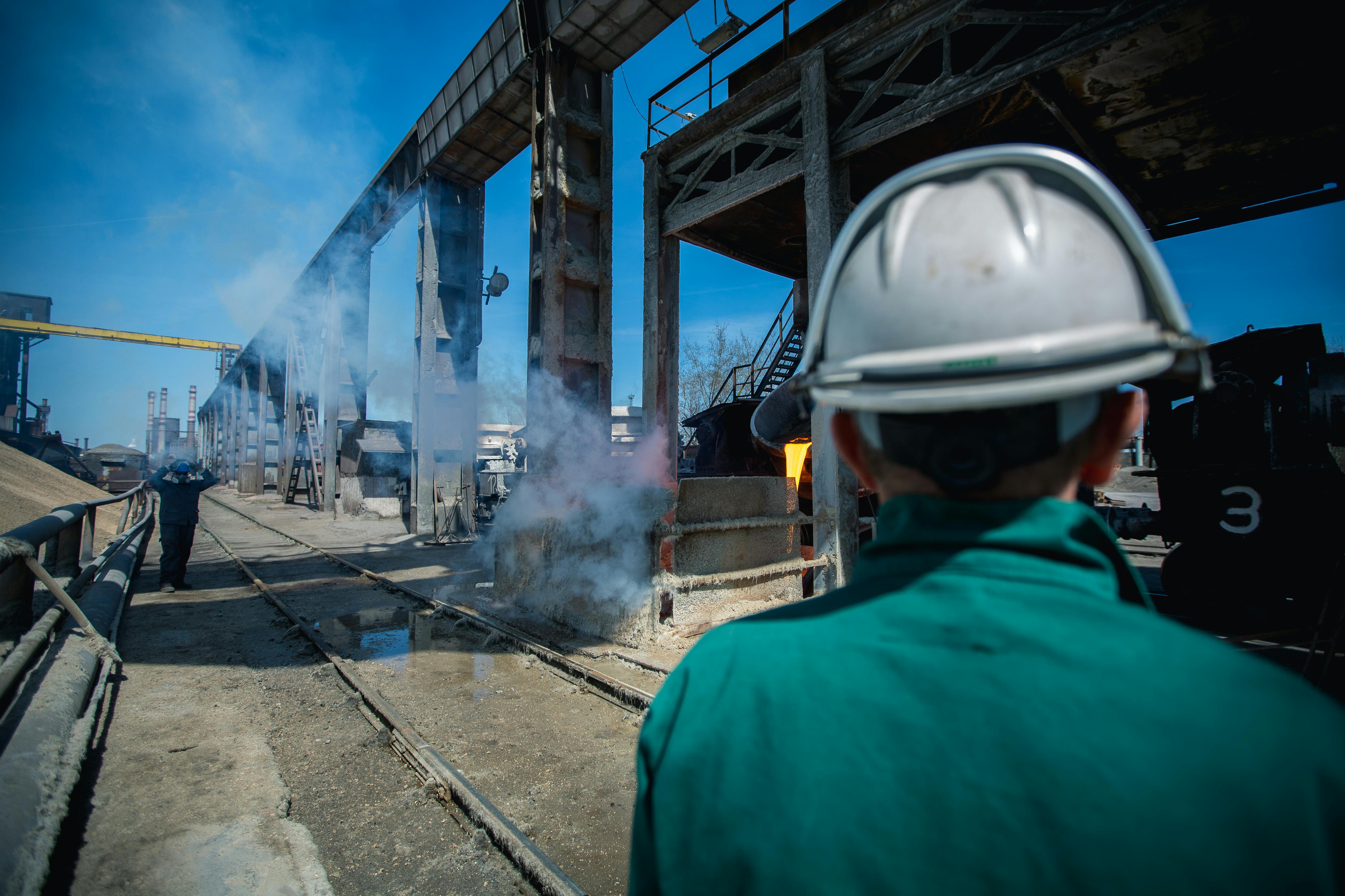 Industrial worker wearing hard hat observing work site with smoke and machinery in a factory setting.
