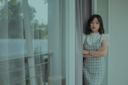 Serious young ethnic woman in stylish dress standing near curtains and window near balcony in daylight and looking at camera with crossed hands