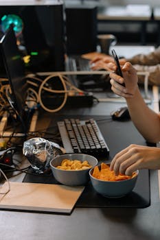 A modern office workspace with snacks, computers, and a person using a phone, capturing a tech-driven lifestyle.