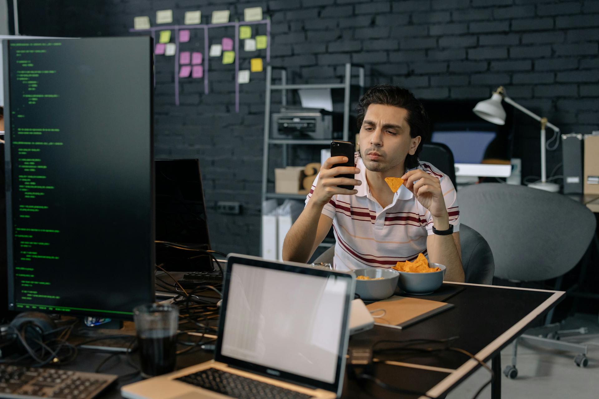 A programmer checks his smartphone while eating snacks at a modern tech office desk.