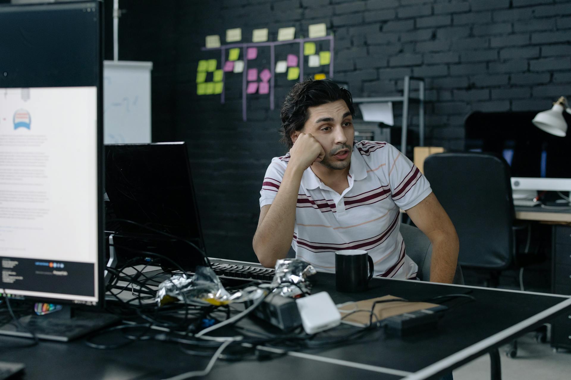 A programmer sits thoughtfully at his desk with computer screens and office setups, reflecting modern workplace dynamics.