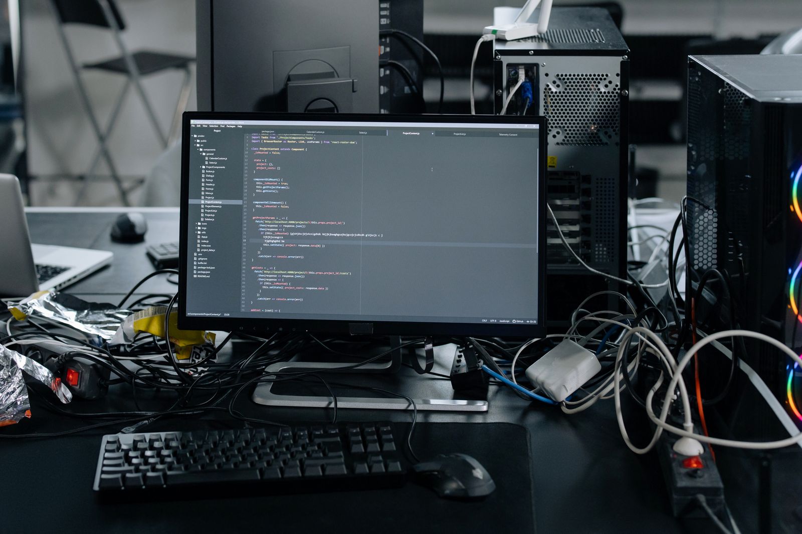 A dark office desk with a widescreen monitor showing lines of code, a mechanical keyboard and mouse in front, a tower case lit with a strip of coloured lights on the right and cables running across the desktop