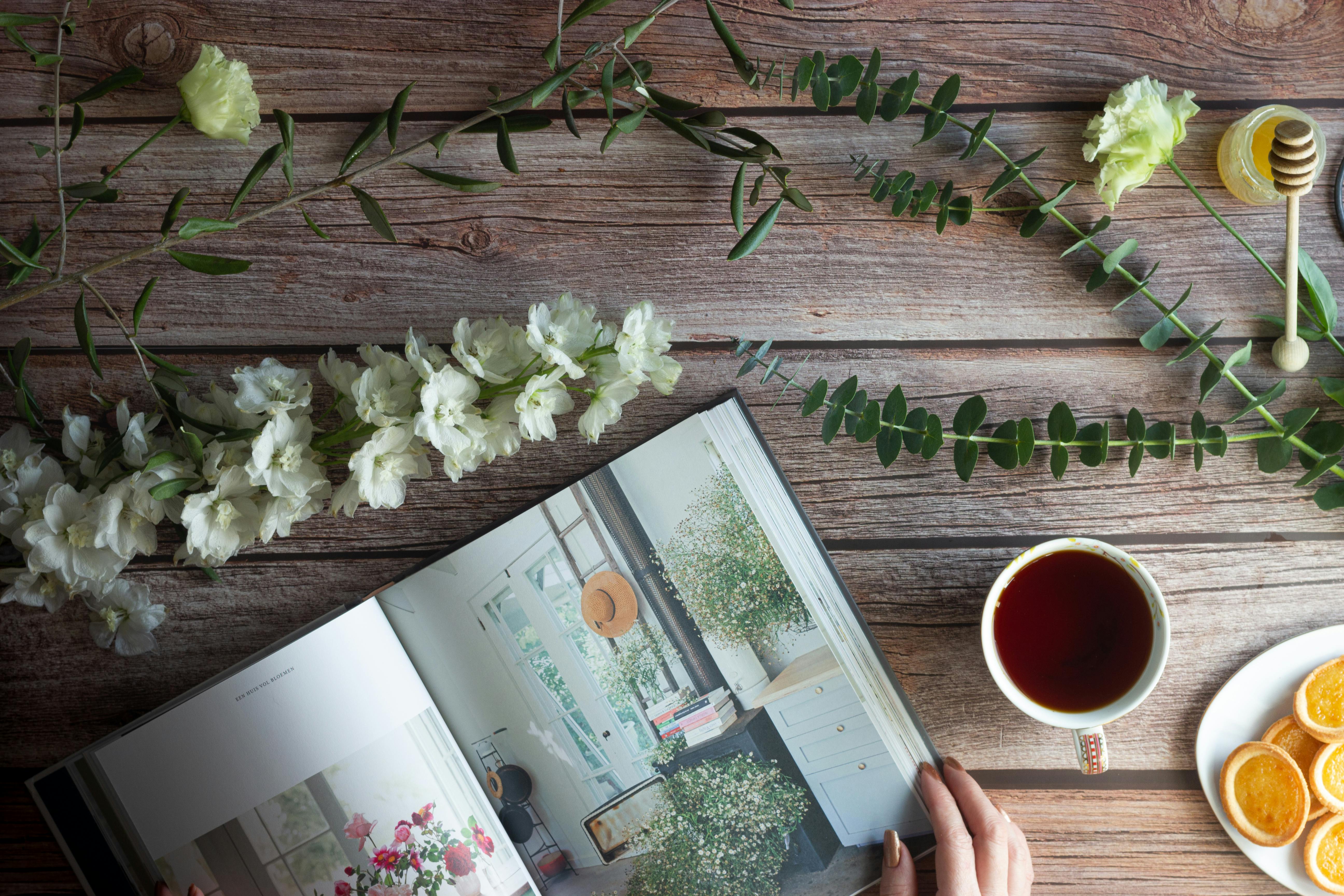 A cozy flat lay featuring a tea cup, open book, flowers, and snacks on a wooden table.