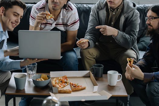 A group of young men enjoy pizza while collaborating on a laptop in a casual office setting.