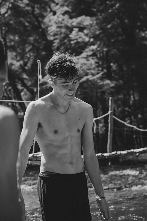 Black and white of positive shirtless male standing in nature against trees and river in sunny day