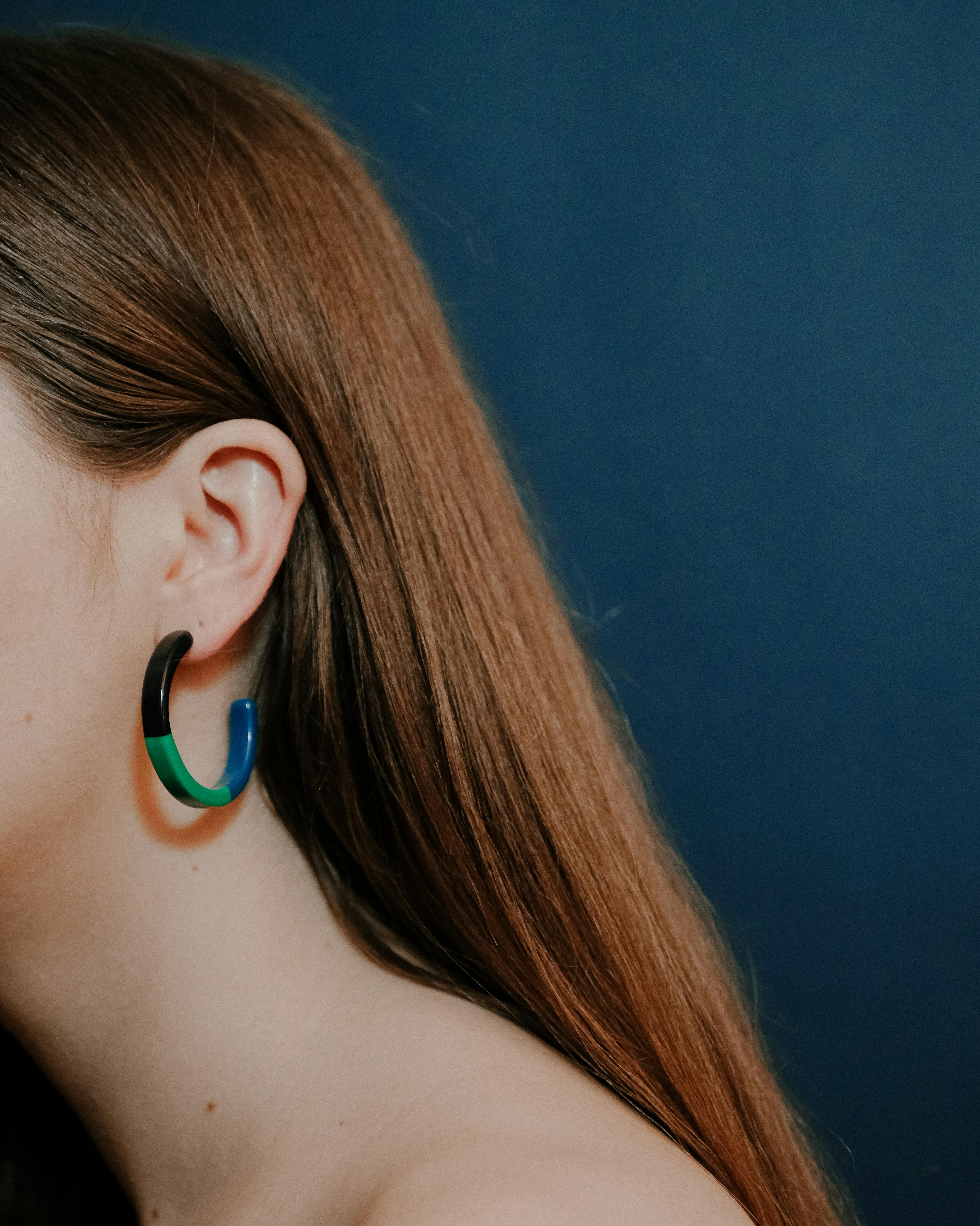 Side view of crop unrecognizable lady with brown hair wearing big hoop earrings with black and green with blue colors on blue background