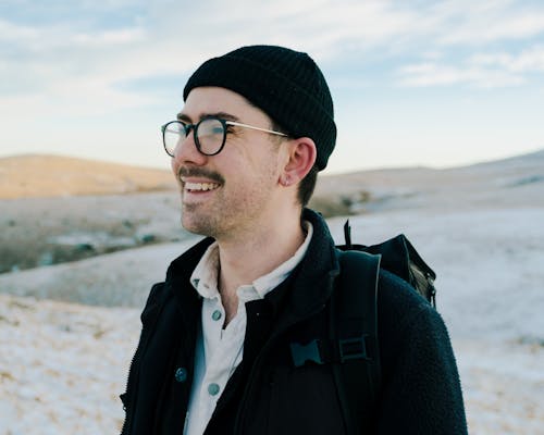 Cheerful male tourist in stylish outfit and eyeglasses standing in snowy hilly valley during trip