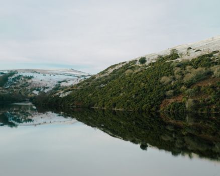 Picturesque view of lake with reflection of grassy slope against snowy hill under cloudy sky